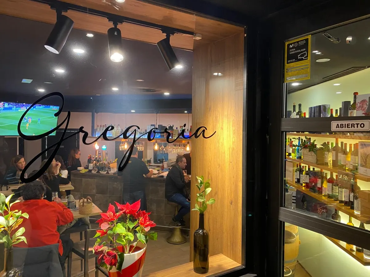 Escaparate de noche con el rótulo caligráfico «Gregoria», la barra al fondo y la vinoteca iluminada a la derecha